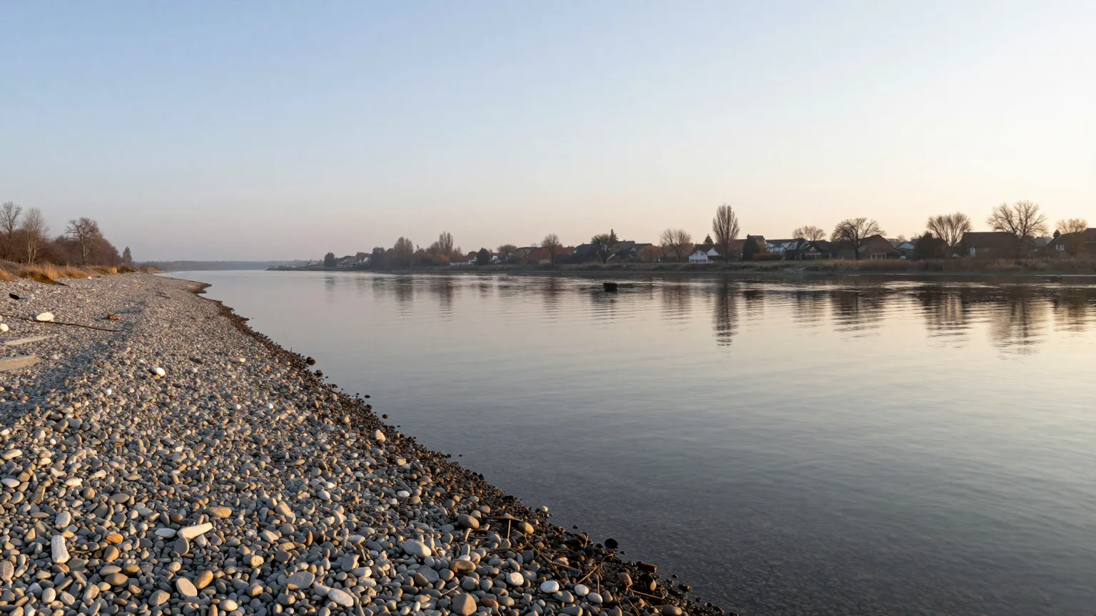 Stilles, weites Wasser an einem Kiesufer in weichem Licht
